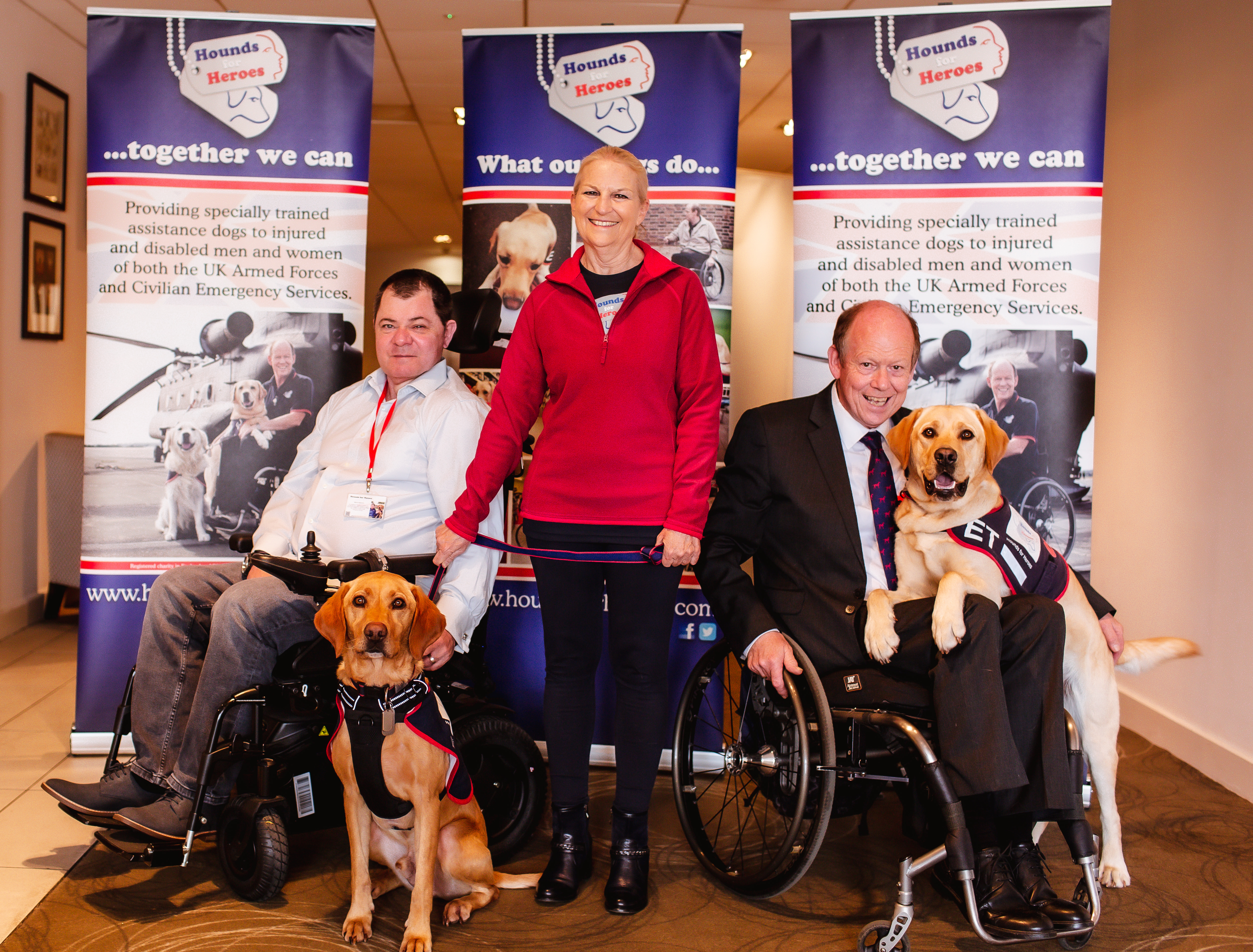 2 Men in wheelchairs with their assistance dogs. Lady stood between them.